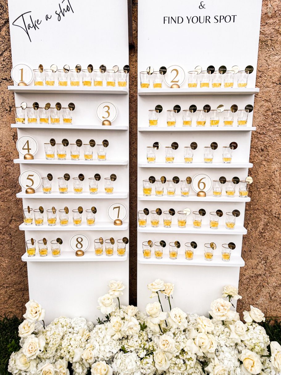 Originelle Hochzeitstischordnung mit Shotgläsern – kreative Idee zur Sitzplatzvergabe, umrahmt von weißen Blumen

